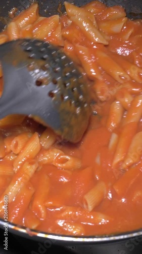 Vertical: A chef expertly tosses pasta with rich tomato sauce in a sizzling pan, blending every strand to perfection for a classic Italian dish served at the catered party event.