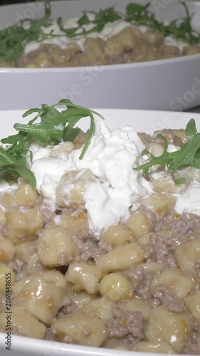 Vertical: Fresh arugula leaves are scattered over trays of gnocchi topped with white ragu sauce and creamy burrata, adding a final peppery touch before serving at the party event.