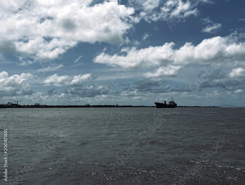 Wide seascape view of dark ocean water under a dramatic blue sky with white clouds, distant cargo ship, shoreline, and calm coastal horizon creating a natural maritime travel background