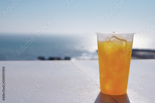 Plastic cup of fresh orange juice with ice cubes on a white table, blurred sea and blue sky background, refreshing summer drink at the beach.