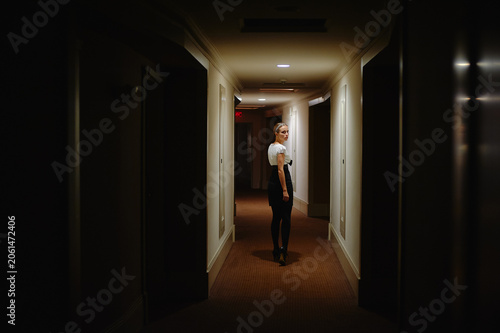 Fotografi A stylish woman in a white blouse and dark skirt stands in a softly lit hotel hallway, turned slightly toward the camera