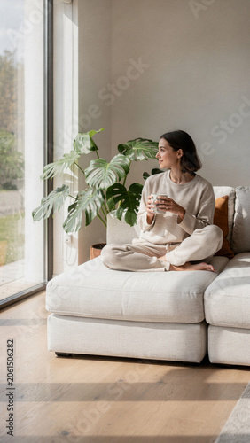 Serene young woman in loungewear sitting on a sofa with a mug, looking out a window in a bright modern living room