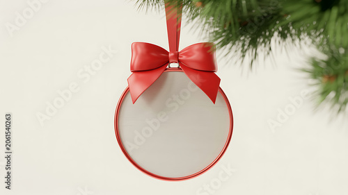 A red metal christmas ornament with a bow hanging on a tree branch with green pine needles on a beige background
