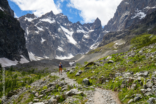 A hiker on a trail through the rugged glacial and rocky landscape of Écrins National Park in the French Alps. Large boulders, alpine vegetation, and scattered larch trees.