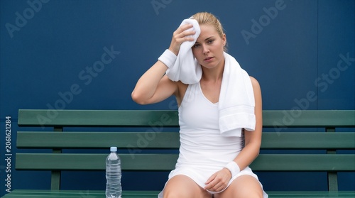 Sweaty tennis player wiping forehead with towel while resting on bench. Athletic woman taking break during intense sports training session.