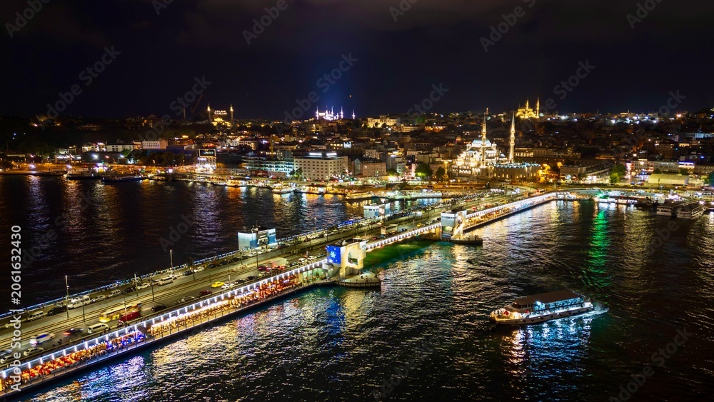 Fototapeta premium Istanbul, Turkey - June 13, 2026: Aerial drone view of Galata Bridge at night, featuring the Golden Horn, bridge lights, waterfront buildings, boats, reflections, and panoramic Istanbul night cityscap
