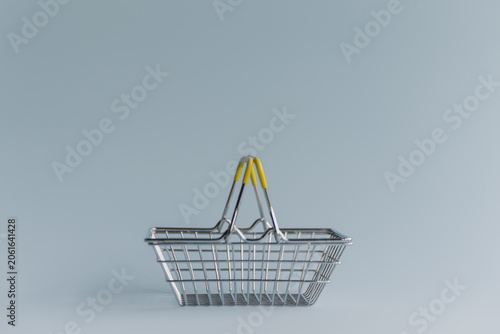 A metal shopping basket with a yellow handle on a light gray background. The basket is empty and has a simple, modern design.