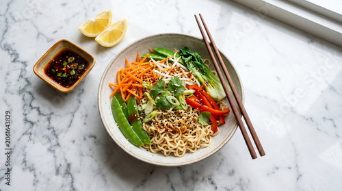 Delicious Asian-style noodle bowl with vegetables and chopsticks on a marble table, served with a side of sauce and lemon wedges for a healthy meal.