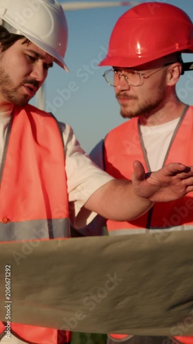 Two professional wind farm workers analyze technical documentation for turbine construction and assembly. Wearing safety helmets and orange vests, they coordinate installation steps and review plans