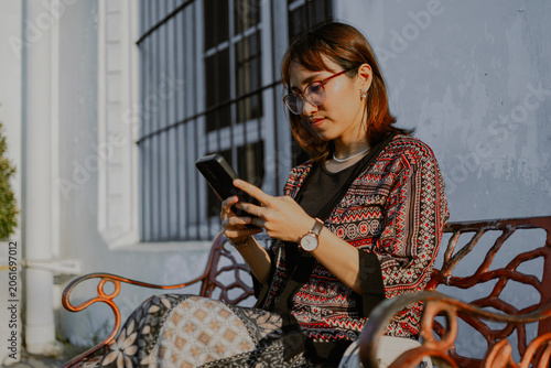 Stylish adult Asian woman in modern batik clothes layered outfit and glasses sits on the bench park using a mobile phone.