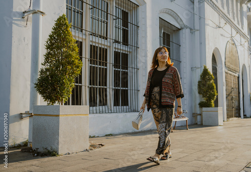 Stylish adult Asian woman in modern batik clothes layered outfit and glasses holds a small bag leather walking in the city.