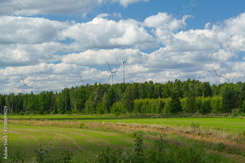 wind turbines behind a forest and agricultural field under a cloudy sky