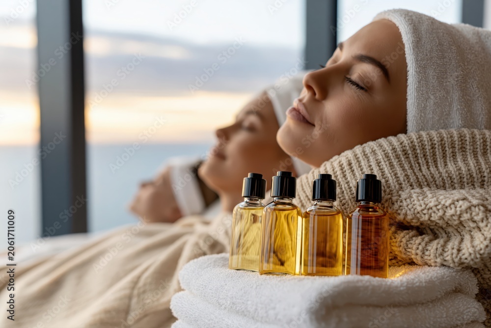 Fototapeta premium Two women relaxing at a spa with essential oil bottles and a serene ocean view at sunset.