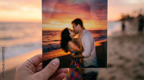 Hand holding photograph of kissing couple on beach during sunset. Romantic memory captured in print. Tropical vacation travel souvenir and intimate relationship keepsake.