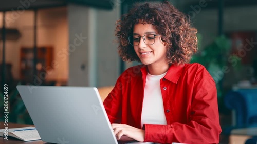 Young indian woman with curly hair and glasses working on laptop computer at desk in modern office or home workspace, wearing red shirt and smiling. Latin or muslim arab student girl studying indoors
