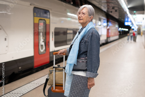 Fotografie Senior woman traveling by train, standing on platform with luggage, anticipating