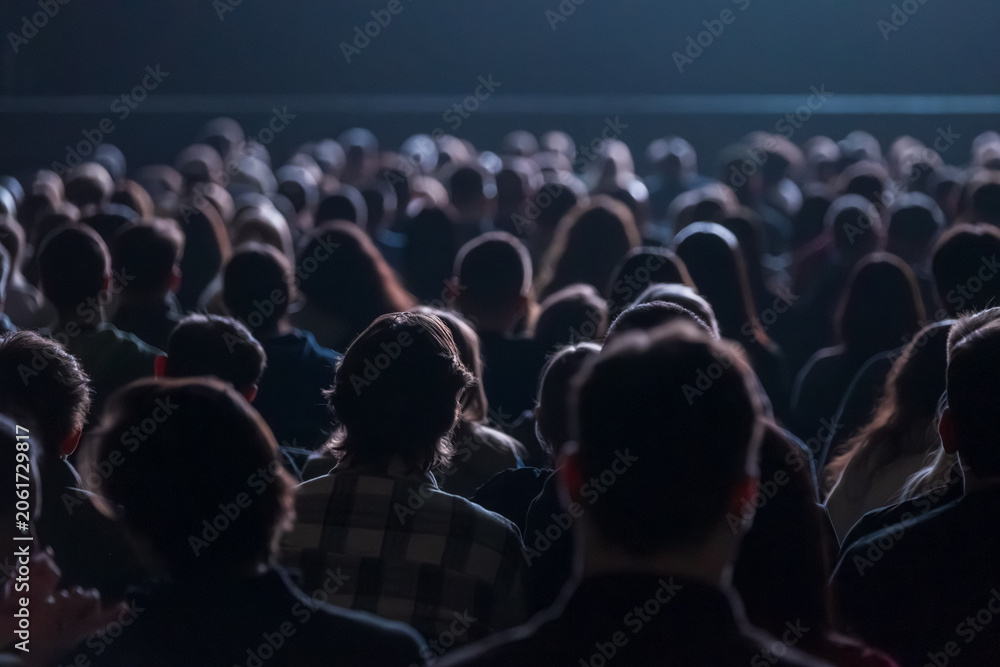 Fototapeta premium Large crowd seated in dim theater, silhouettes of heads filling rows, atmosphere of anticipation and focus.