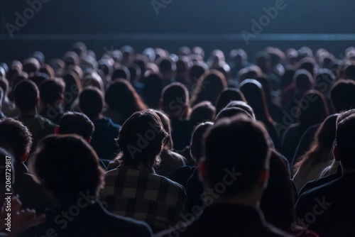 Large crowd seated in dim theater, silhouettes of heads filling rows, atmosphere of anticipation and focus.