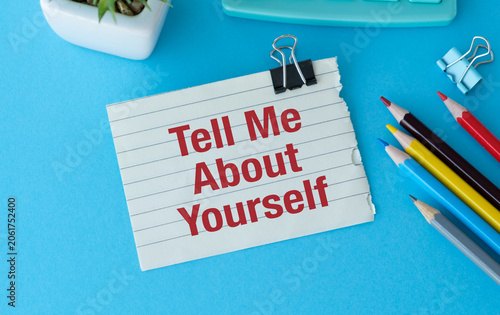 Bright red text on lined paper reads Tell Me About Yourself. Paper clipped to a vibrant blue surface with colorful pencils nearby.