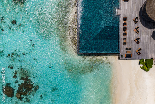 Aerial view of a Maldives luxury resort shows an infinity pool by a pale deck, a white sand beach, a turquoise lagoon with coral heads, and a thatched roof at midday.