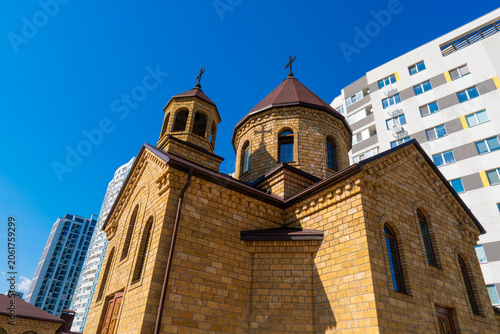 Armenian church built of yellow tuff stone on a sunny day. A traditional Christian temple contrasts with modern high-rise buildings under a blue sky