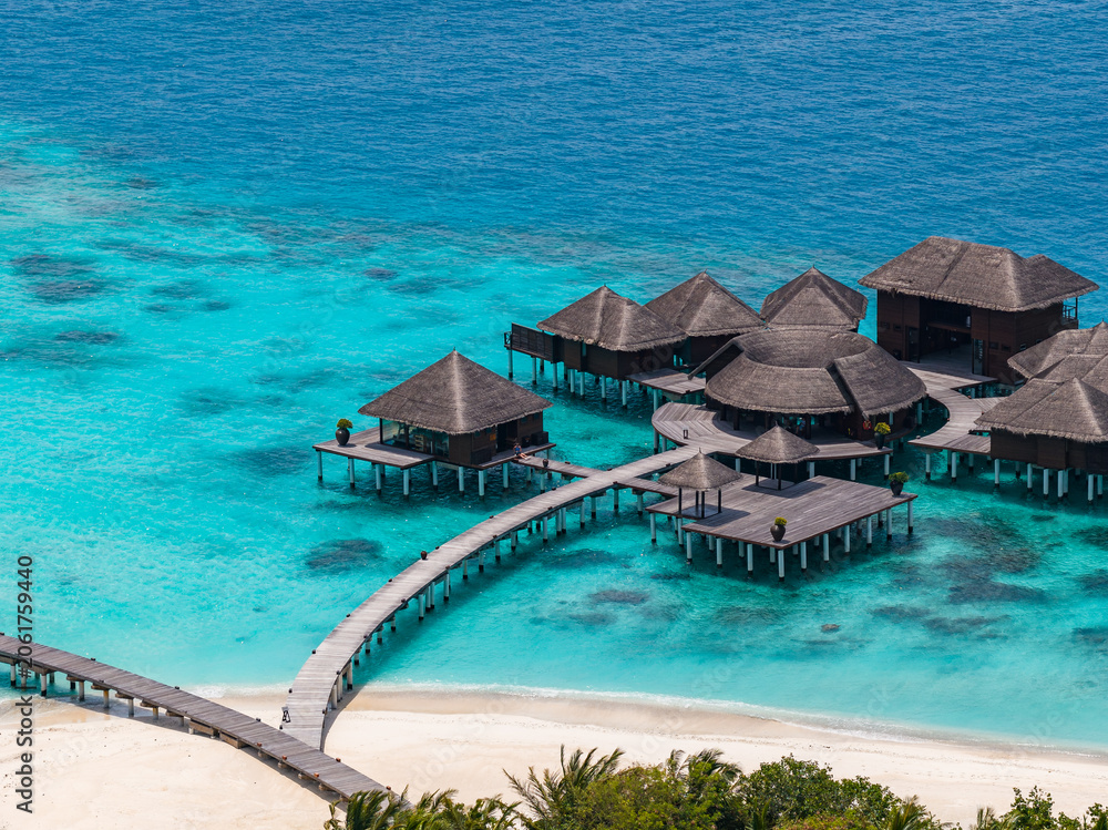 Fototapeta premium Aerial view of a Maldives atoll resort shows a curving boardwalk, thatched overwater villas, white sand, and lagoon gradients under midday sun and clear skies.
