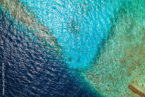 Bild auf Leinwand Aerial view of a Maldives atoll shows deep navy ocean meeting turquoise shallows over a coral reef, with rippled water, bright sunlight, and a small wooden jetty at lower right