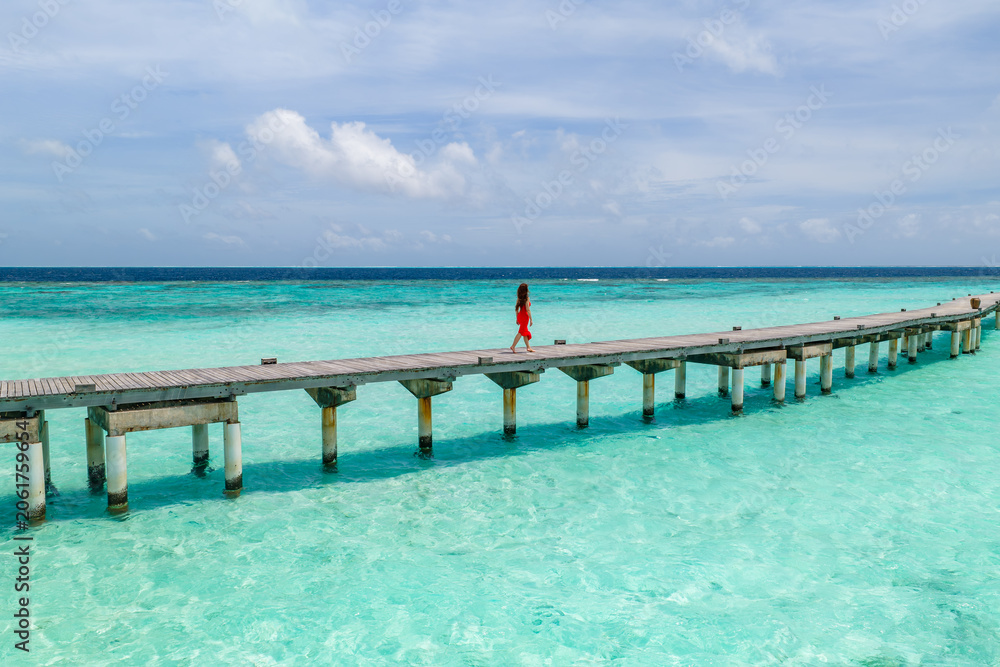 Fototapeta premium A wooden jetty in the Maldives extends over turquoise shallows to deep water, with one person in a bright red dress walking, under midday sun and soft clouds.