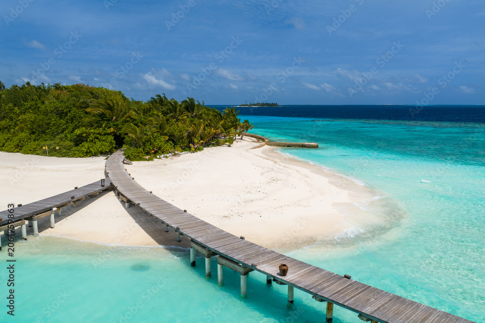 Fototapeta premium Aerial view of a Maldives resort island with a wooden boardwalk over turquoise lagoons, a crescent of white sand, green palms, and a jetty in bright daylight.