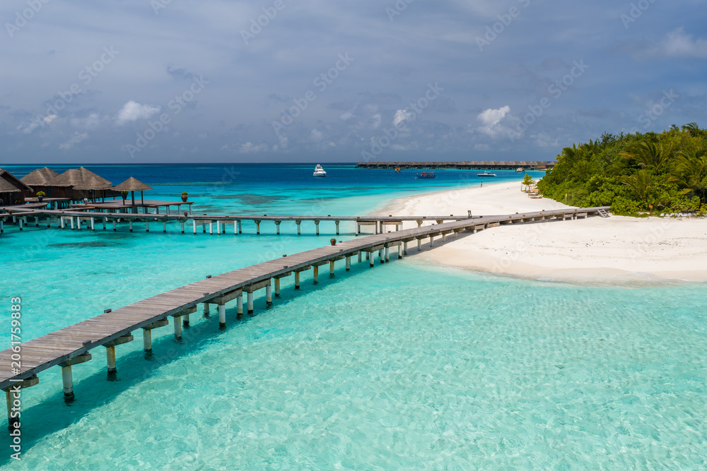 Fototapeta premium Midday Maldives resort scene shows S curve pier over turquoise shallows to overwater villas by a white sand lagoon, palms on the isle, and a small boat on the horizon.