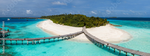 Obraz na plátně Aerial view of a Maldives resort island shows a crescent sand beach, palm forest, curving boardwalks, overwater villas, jetties, a distant pier, and a boat under midday light