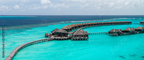 Foto Aerial Maldives resort shows crescent boardwalk of overwater villas, thatched roofs, infinity pool terraces, shallow reef shelf, and deeper Indian Ocean under midday sun
