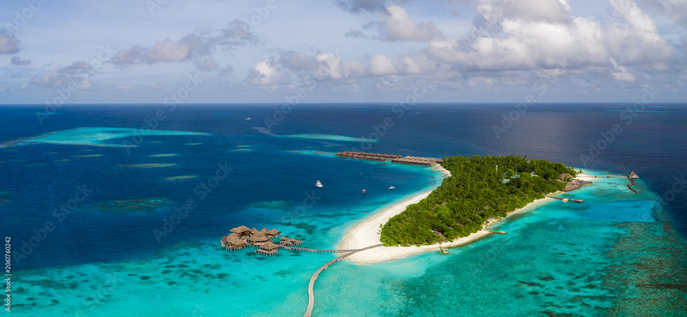 Obraz premium Aerial scene of a Maldives resort island with white sand, turquoise shallows, overwater villas on a curved pier, wooden pavilions, jetty, palms, and midday light.
