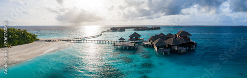 Photos Aerial view of a Maldives resort, a sinuous boardwalk links overwater villas, a white sand beach and palm island sit left, golden hour light reflects across the lagoon