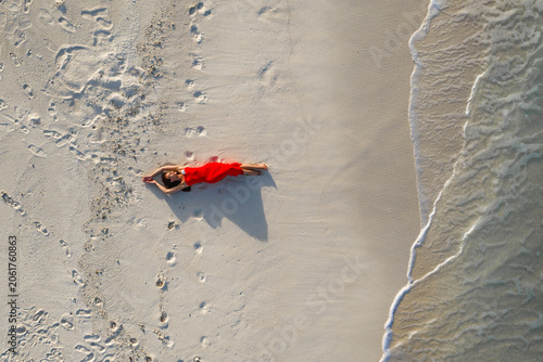 Aerial view of Maldives beach shows a woman in a red dress on white sand, footprints leading to her, gentle shoreline ripples, long shadow, golden hour minimalism.