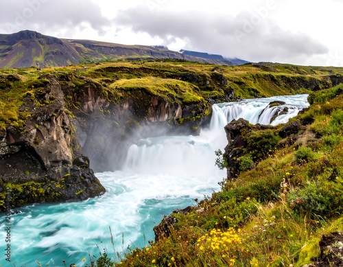 A serene waterfall cascades into a vibrant blue river surrounded by lush greenery and rugged rocky cliffs under a cloudy sky