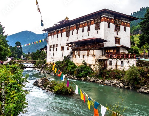 A serene white building with brown accents and a tiled roof situated beside a flowing river with prayer flags