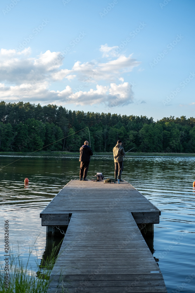 Fototapeta premium Two male friends enjoying a peaceful afternoon fishing from a wooden pier on a calm lake bordered by a dense pine forest under a blue sky.