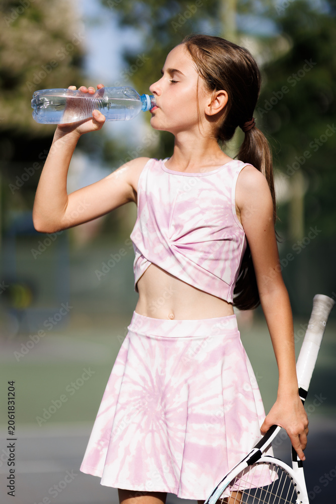 Naklejka premium Young girl drinks water while taking a break from tennis practice on a sunny day at the local tennis court