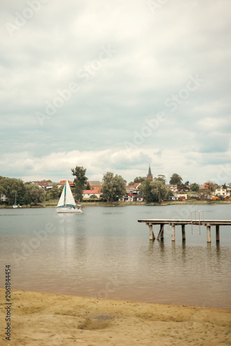 Wooden pier extending into a peaceful lake with a sailing yacht gliding across the water in the background