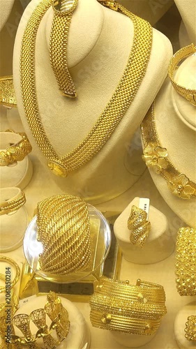 Close-up of ornate gold jewelry displayed in a shop window, showing necklaces, bracelets, rings and bangles with a slight camera shift across the luxury showcase