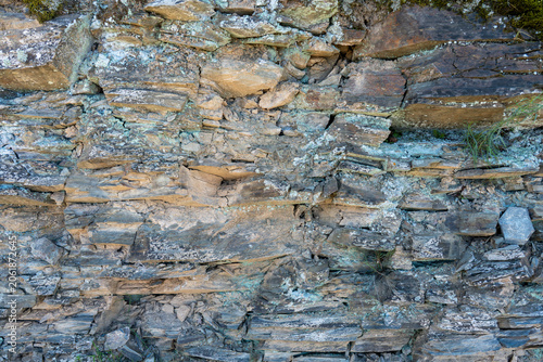Photos Full-frame texture of a crumbling geological rock face, displaying earthy brown and grey tones, sharp fractures, and subtle traces of dry grass and moss
