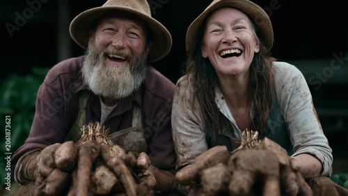 Elderly couple laughing while holding muddy yams and root vegetables on rustic organic farm porch during daytime harvest