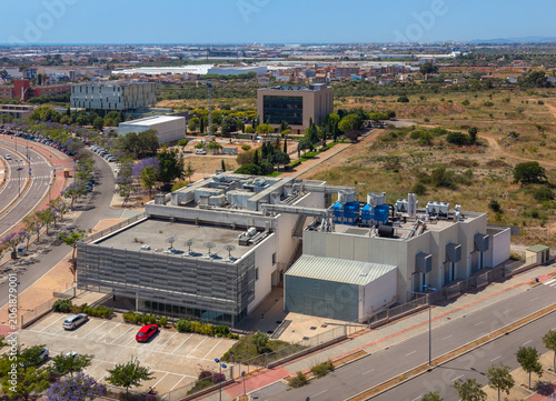 Aerial overhead view of a data centre facility with rooftop cooling towers, blue chillers and satellite dishes surrounded by blooming jacaranda trees in a technology park, Spain