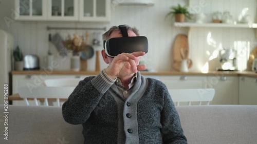 Excited mature senior man wearing using virtual reality metaverse VR glasses headset at home. Grandfather touching air during VR experience on virtual reality helmet. Simulation hi-tech videogame