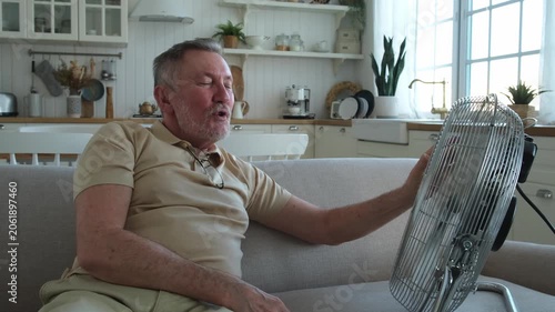 Senior man refreshing himself near fan during summer heat wave hot day. Elderly man suffering from summer heat trying to survive with electric fan. Man enjoying refreshing air blowing from ventilator