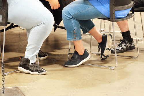 Seated legs and feet under a table at a casual group gathering showing assorted sneakers, socks and denim pants in a cropped candid view with folding chairs on a concrete floor, social interaction.