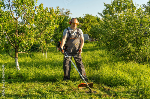 Fotografía A worker stands in a garden, trimming grass with a grass trimmer on a sunny day