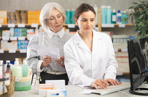 фотографія Woman employee in labcoat works in pharmacy using computer and collaborate with senior female client