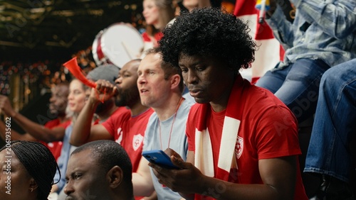 Foto Black sports fan placing football betting wager through mobile sportsbook during live soccer match with cheering supporters at the stadium, man experiencing sports betting tension and anticipation